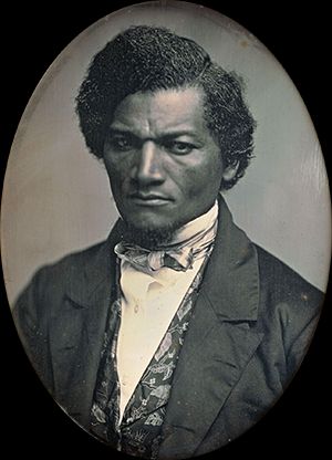Oval-framed daguerreotype portrait of Frederick Douglass, a prominent 19th-century African American abolitionist, writer, and orator. He wears a dark jacket, patterned waistcoat, white shirt, and bow tie. His expression is serious and resolute, with his wavy hair parted to the side. The photograph reflects his dignity and strength as a leader in the fight against slavery.