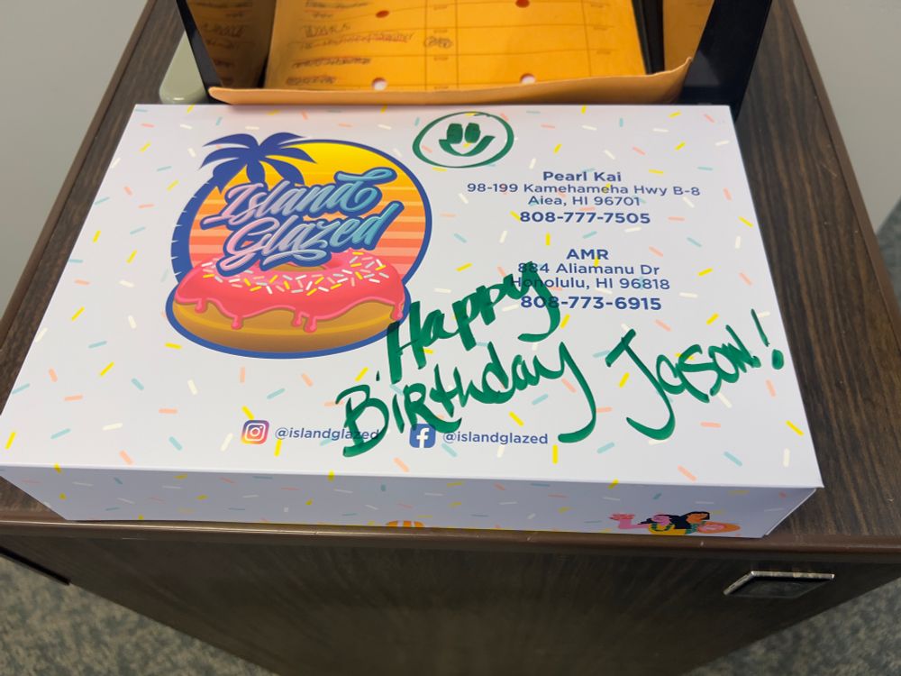 A white rectangular box containing half a dozen donuts with the logo for “Island Glazed” with the name over a sunset and palm tree with a strawberry sprinkle glazed donut underneath.  To the right is the address for two of its location, at Pearl Kai in Aiea, Hawaii and AMR in Honolulu, Hawaii.  On the top center is a smiley face and on the bottom is a message that reads “Happy Birthday Jason!” written with a green marker.
