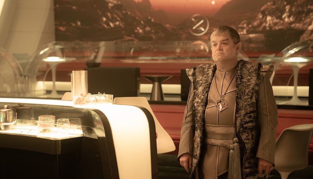 Doug (played by Patton Oswalt), a Vulcan, dressed in traditional Vulcan clothing and standing in the port galley of the USS Enterprise, as he sees his ex, Una Chin-Riley, accompanied by Spock (both off screen in this image).  From “Star Trek: Strange New Worlds” season 3, episode 8, “Four and a Half Vulcans.”