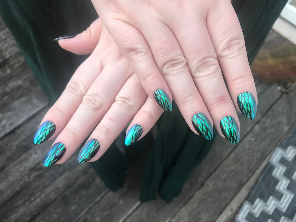 Two hands laid overtop one another to display nail art. Shimmery green-blue nails with black line-work detailing to evoke the image of beetle wings!