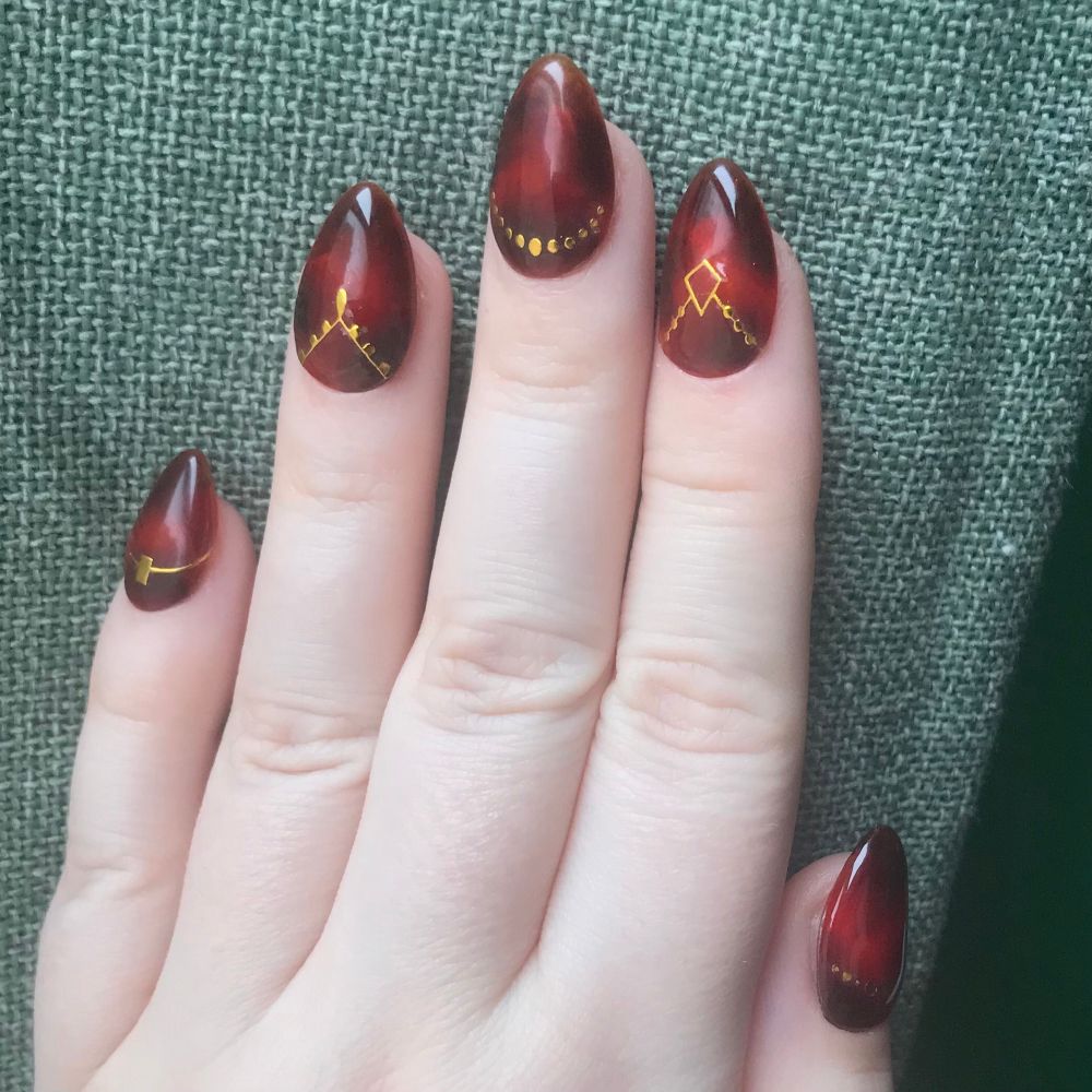 A hand is posed against a green curtain to display nail art. The design is tortoise shell with gold decal stickers near the cuticle line.