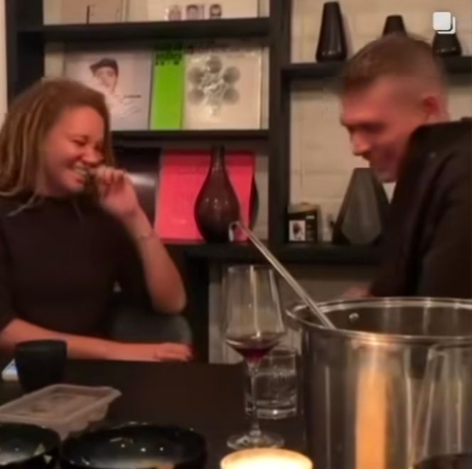 A picture of Ayoola Smart, a redheaded woman with tan skin, and Josha Stradowski, a redheaded man with tan skin, sitting on opposite sides of a table with some partly filled stemware, partly filled bowls, a candle, and a steel pot on it. They are both laughing heartily as if over some privately shared hilarity.
