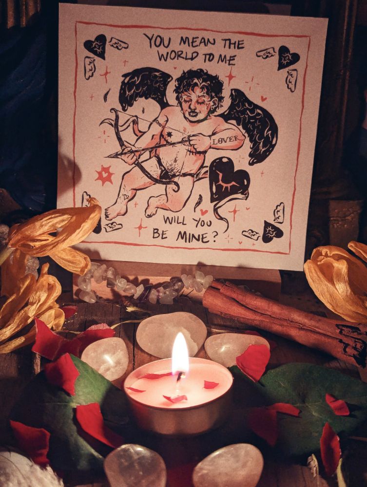 A lit candle is surrounded by rose petals, flowers, leaves, and crystals. In the background is an art print of a tattooed cherub that reads YOU MEAN THE WORLD TO ME... WILL YOU BE MINE?