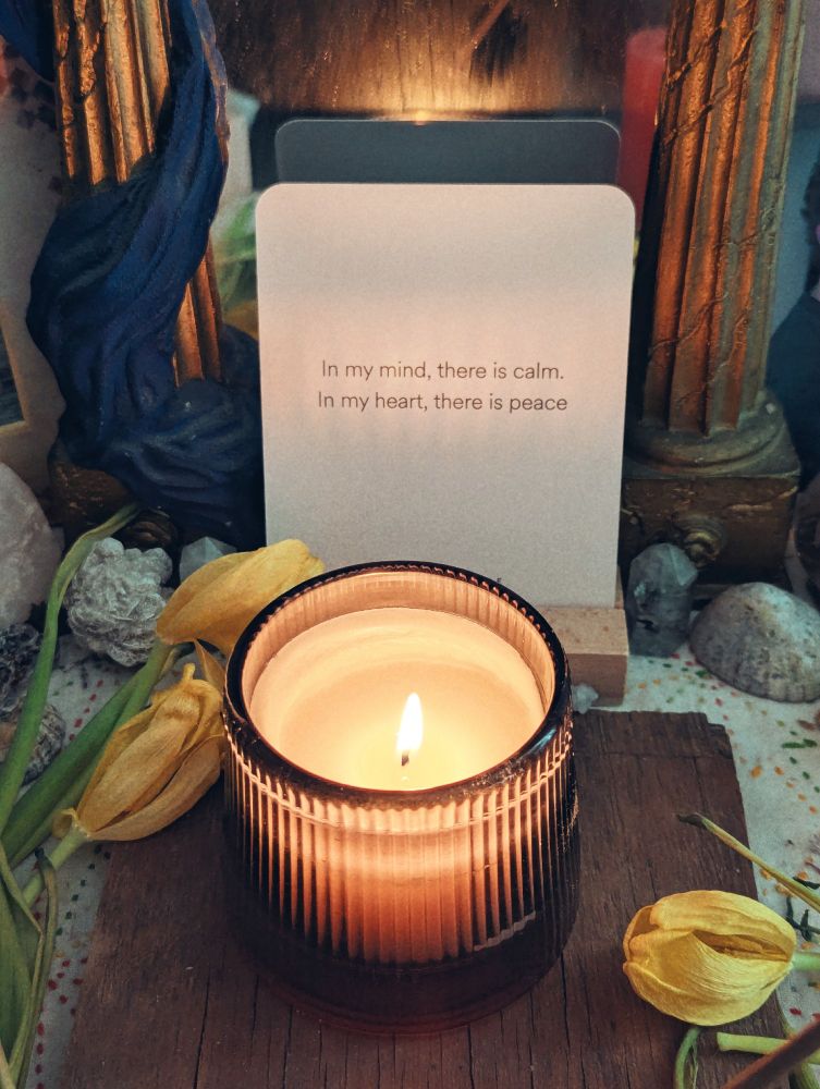 A candle on an altar, surrounded by flowers and crystals and shells. There's a mirror framed by golden pillars, and resting against the mirror an affirmation card: In my mind, there is calm. In my heart, there is peace. 