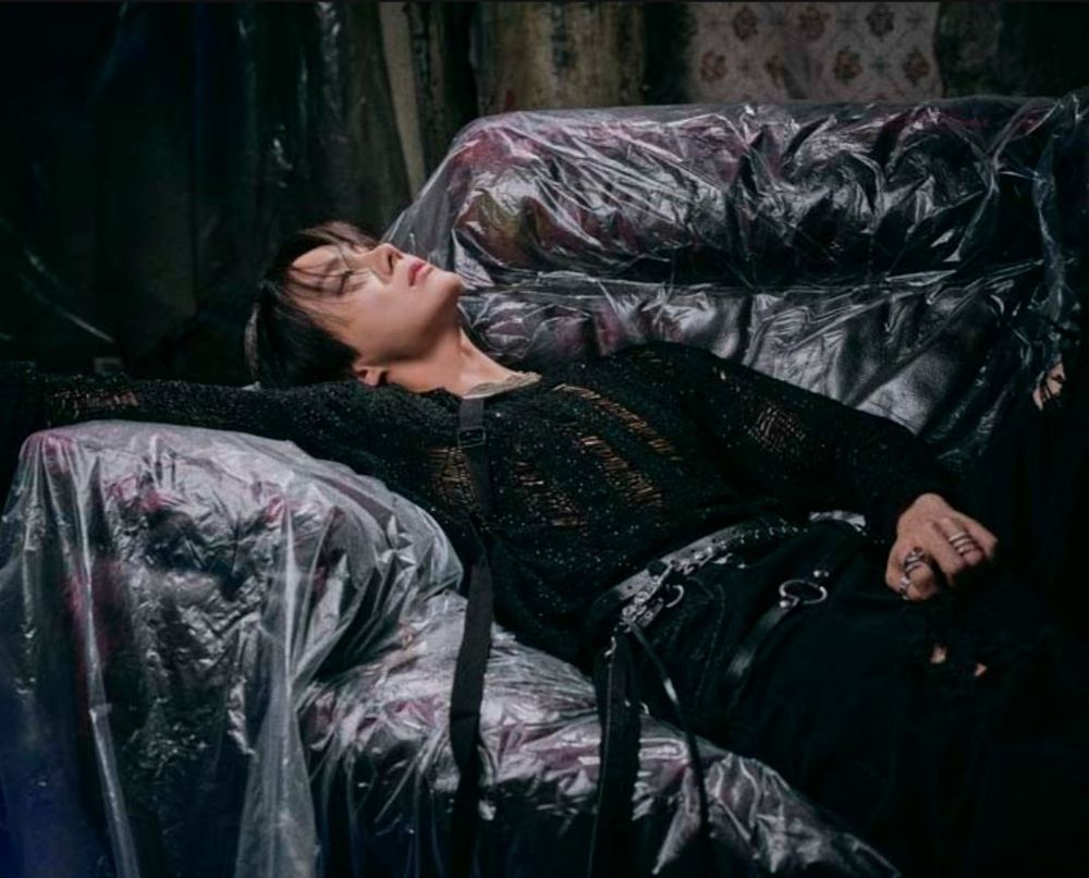 hoseok lying on a couch that is helpfully covered with plastic. he is looking up, grabbing his crotch like a little hoe, and wearing a ripped up black sweater with baggy black pants and lots of strappy belts