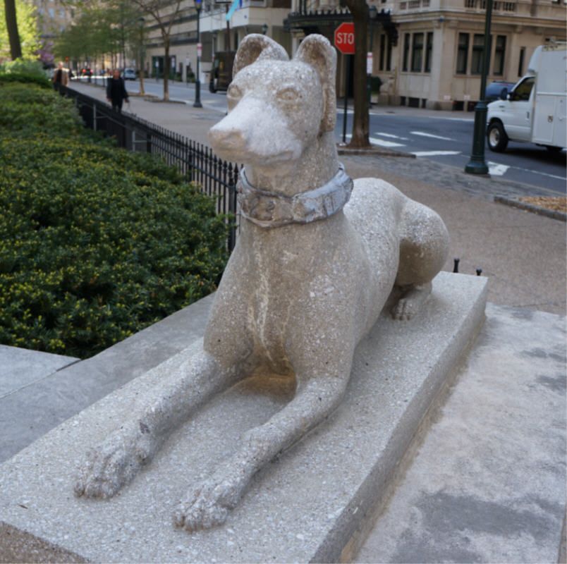 life-size or slightly larger stone or concrete statue of a crouching greyhound on top of an entrance pillar, taken from the front and slightly to the left.  The dog is staring straight ahead, ears up, front legs extended, with the back legs hunched.  There is a collar around the neck.  Also visible in the picture, green bushes, a black iron fence, trees, people walking, sidewalk, street, a white truck, buildings, and a stop sign.  An identical dog on the opposite side of the walk is not pictured.