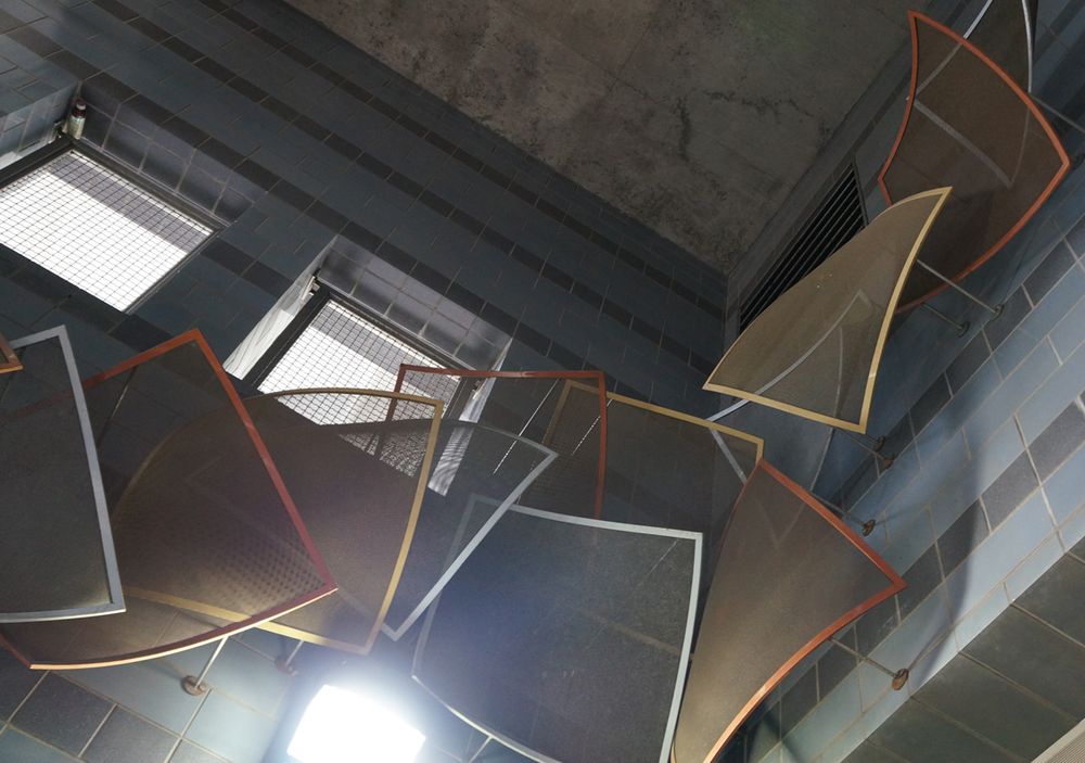Different colored (red, yellow, and blueish grey) bent metal squares about a foot or two on a side with thin solid borders and mesh interiors  rise up the stairwell to the ceiling like abstract leaves blown by the wind.  Also visible in the picture are the stairwell ceiling, windows, and tiled walls, and the rods attaching the squares to the walls.  The bluish grey, while not a standard autumn leaf color, matches the lighter wall tiles.
