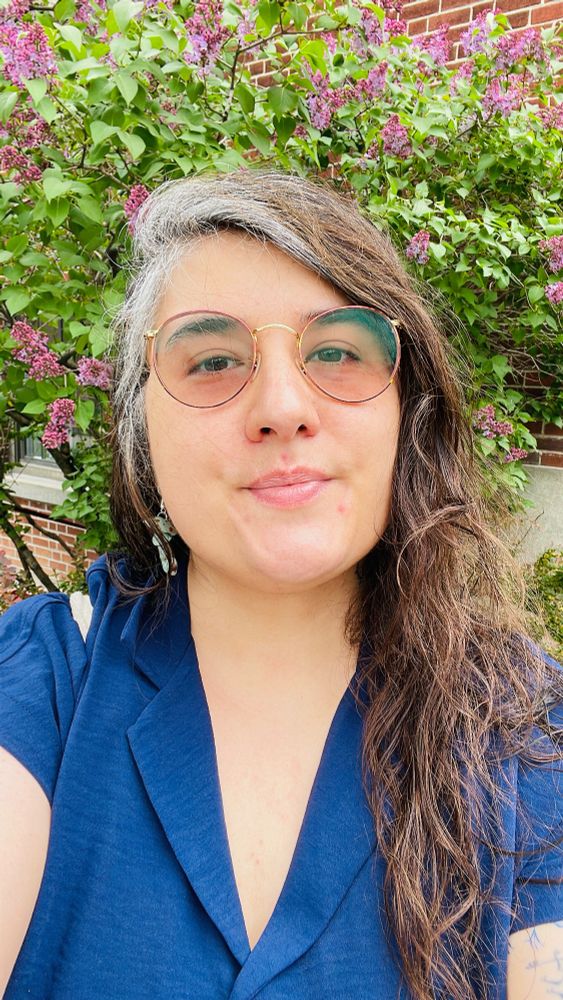 Photo of P.Andi below a lilac bush starting to bloom. She has a blue shirt on. They have a white streak of hair reminiscent of Rogue from X Men and long brown hair.
