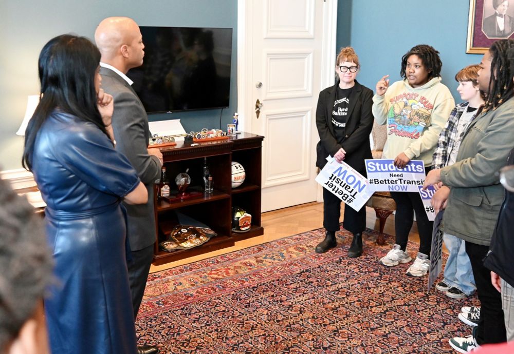 A photo of students and educators sharing their stories and describing their need for better transit in Baltimore with Governor Wes Moore and Lieutenant Governor Aruna Miller. Some of the students and educators are holding #BetterTransitNOW rally signs.