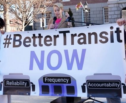 Holly Arnold, administrator of the MTA, shares her vision about the future of transit in Baltimore at a makeshift podium outside the Maryland State House. She stands in front of a large sign that reads "#BetterTransitNOW" with the words "Reliability", "Frequency", and "Accountability" over drawings of buses and trains.