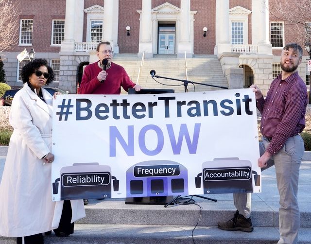 Three organizers stand outside the Maryland State House holding a large banner that reads '#BetterTransitNOW' in bold letters. Below, the words 'Reliability,' 'Frequency,' and 'Accountability' are displayed over stylized images of buses and a train. Paul Sturm, the lead campaign organizer, speaks into a microphone while the other two organizers hold the banner, advocating for improved public transit.