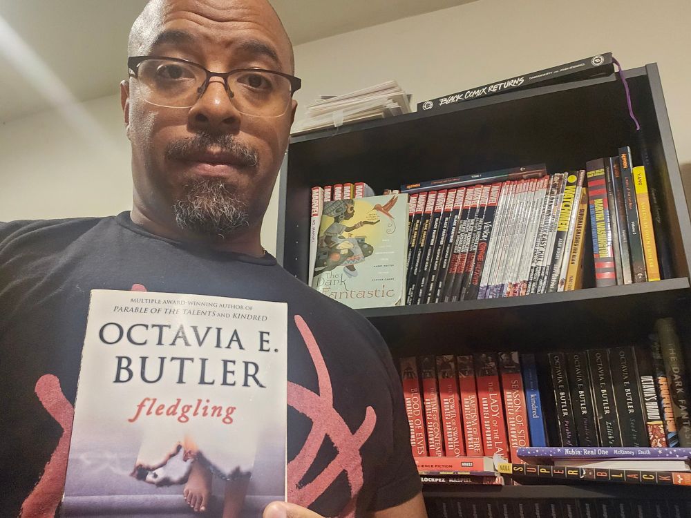 A man standing in front of a bookshelf while holding a book, _Fledgling_ by Octavia Butler
