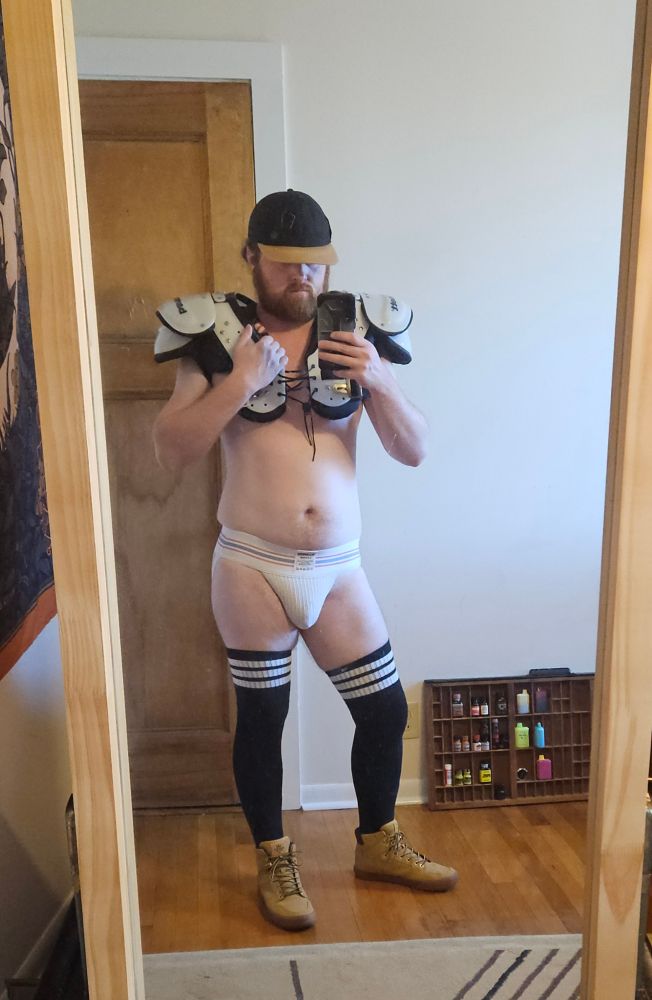 Mirror pic of a male, front view, wearing boots and black thigh high socks, a jockstrap, football shoulder pads, and a baseball cap with brim covering his eyes.