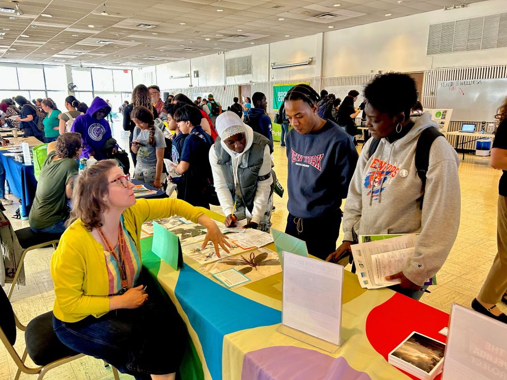 Students gather around display tables in a convention setting to learn about projects being undertaken by cultural institutions like the New York Botanical Garden.