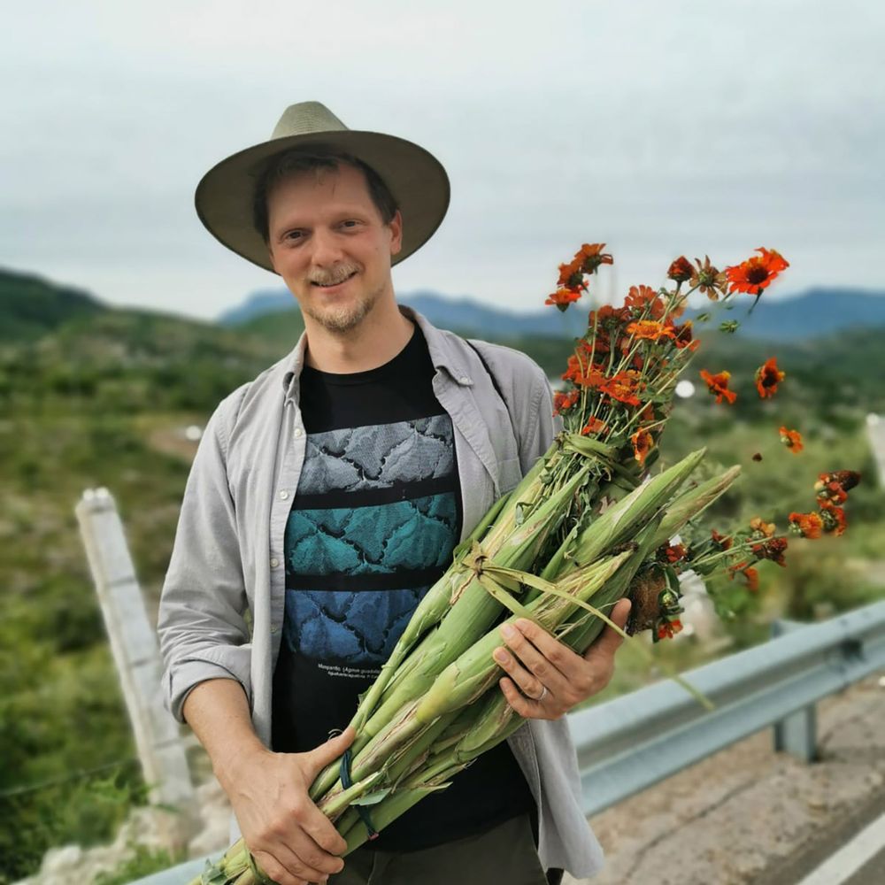 A person in a brimmed hat and gray shirt poses for a photo outdoors while holding a sheaf of green vegetables with orange flowers