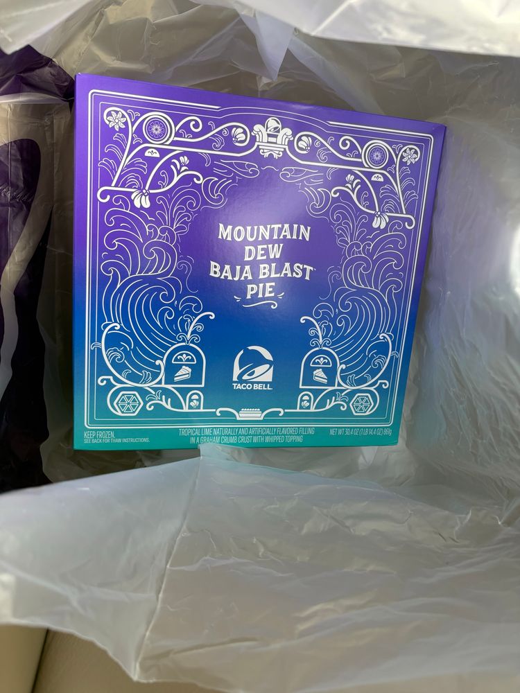 A box containing the Mountain Dew Baja Blast Pie. The box is within a bag. The box has patterns and design of what looks like waves and some Thanksgiving theming. It is a gradient going from purple to blue to teal.