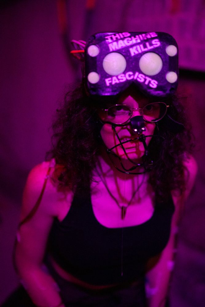 amber as a mech pilot, with coveralls unzipped to her waist showing a sports bra and bare midriff, she has iv tubes in her arms, and a headset on her head with stickers reading "KEEP MUZZLED", "C4-621", and "THIS MACHINE KILLS FASCISTS." she's also wearing a muzzle