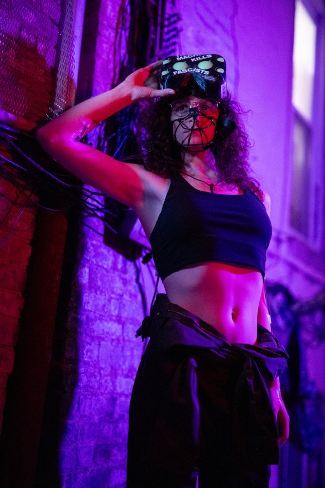 amber as a mech pilot, with coveralls unzipped to her waist showing a sports bra and bare midriff, she has iv tubes in her arms, and a headset on her head with stickers reading "KEEP MUZZLED", "C4-621", and "THIS MACHINE KILLS FASCISTS." she's also wearing a muzzle