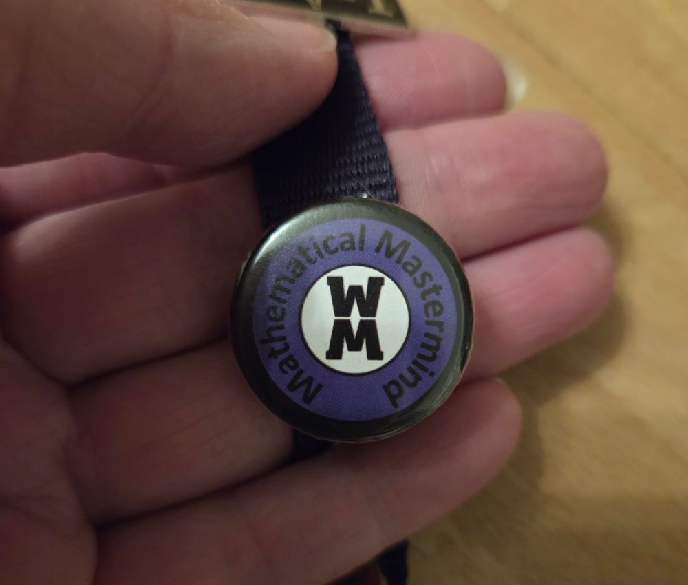 A round badge with a black rim, a purple outer circle with "Mathematical Mastermind" around the outside, and a white centre with the letters W and M. The badge is about 2cm in diameter.