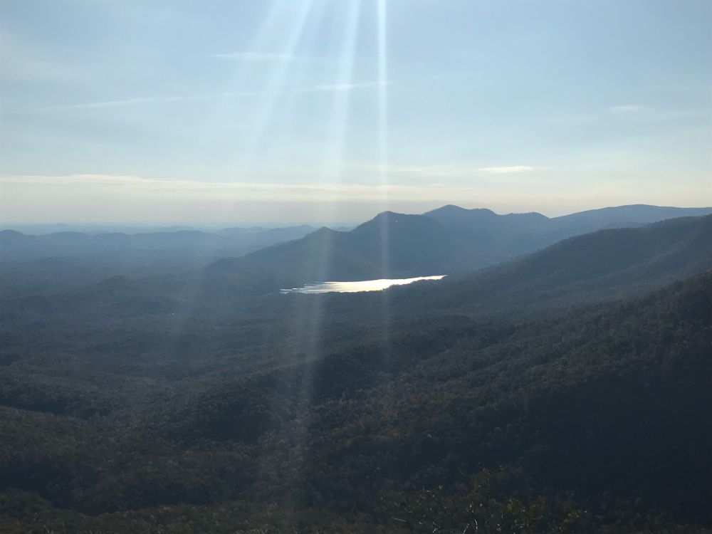 View of blue ridge mountains, taken atop a gneiss. A lake can be seen in the center of the frame, reflecting the sun. Sunbeams come from the top of the frame. Clouds recede into the distance. 