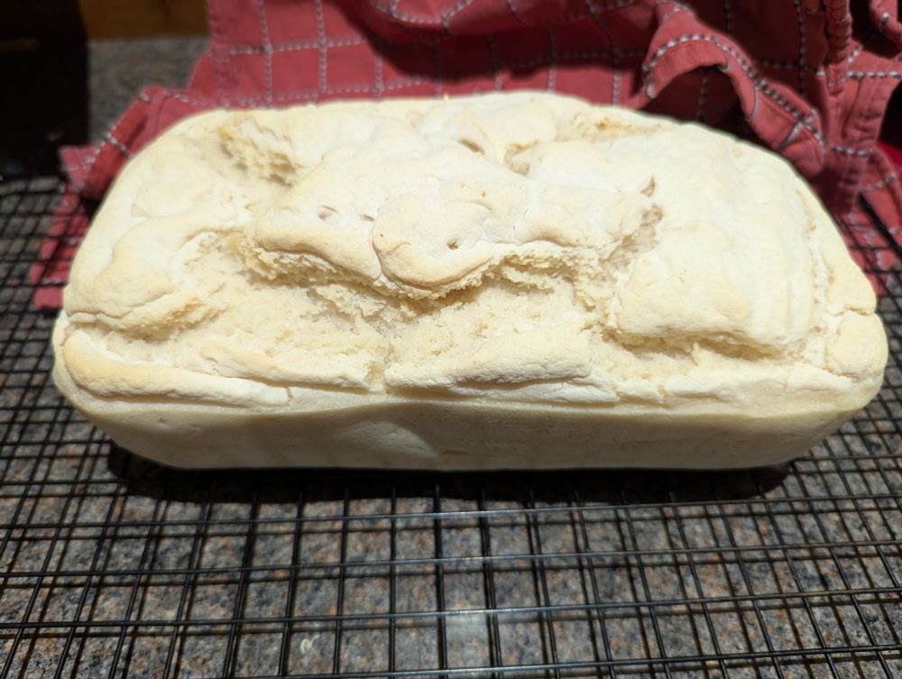 A fresh loaf of sourdough bread, with a pale crust, on a cooling rack. The fluffy internal structure is poking through the top.