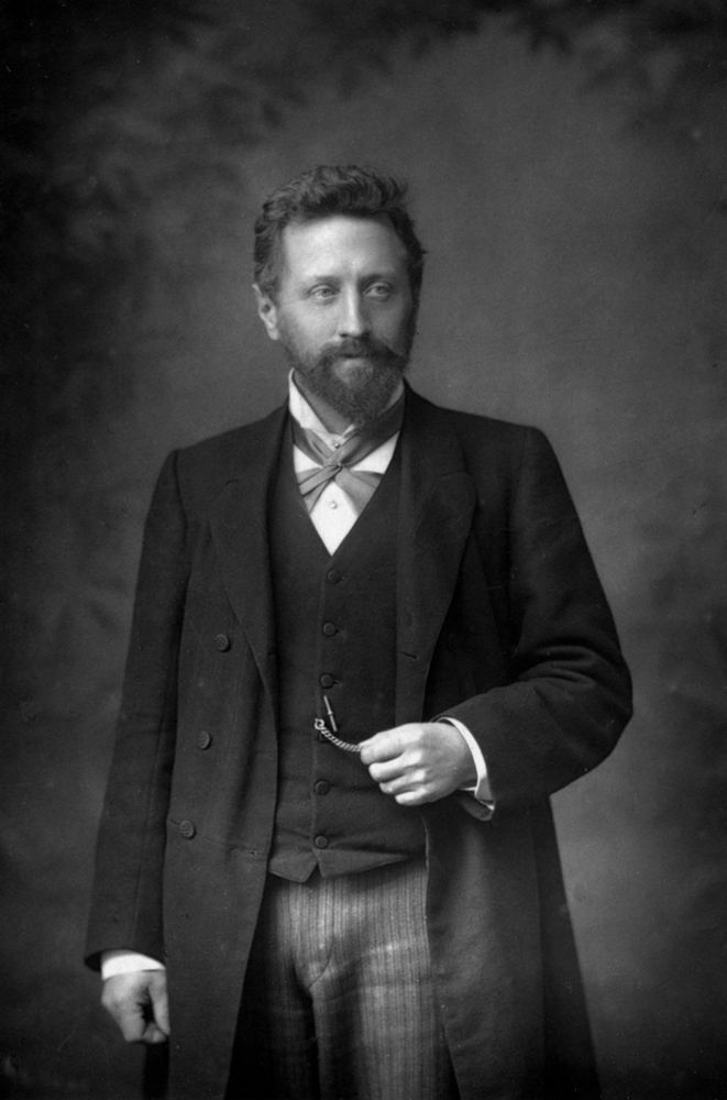 Profesor William Edward Ayrton en 1893. Retrato fotográfico del profesor donde aparece con el puño derecho cerrado hacia abajo y la mano izquierda sujetando un reloj. Tiene el rostro afilado con barba completa y pelo corto. 