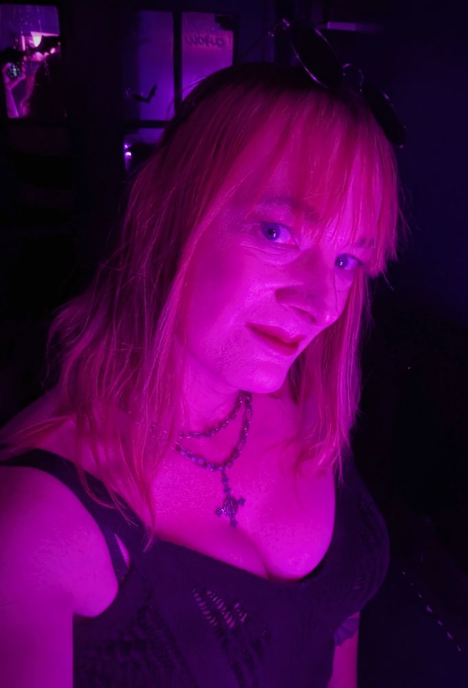 Me in neon pink lighting wearing a black knitted crop top with a cross necklace hanging between my cleavage.