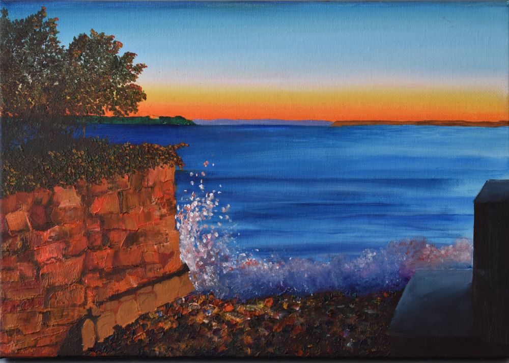 Pebbled lane with stone wall on left and concrete steps on right. High tide and sea splashing on the pebbles. It's dawn with an orange sky along the horizon, the light is catching the spray of the water.