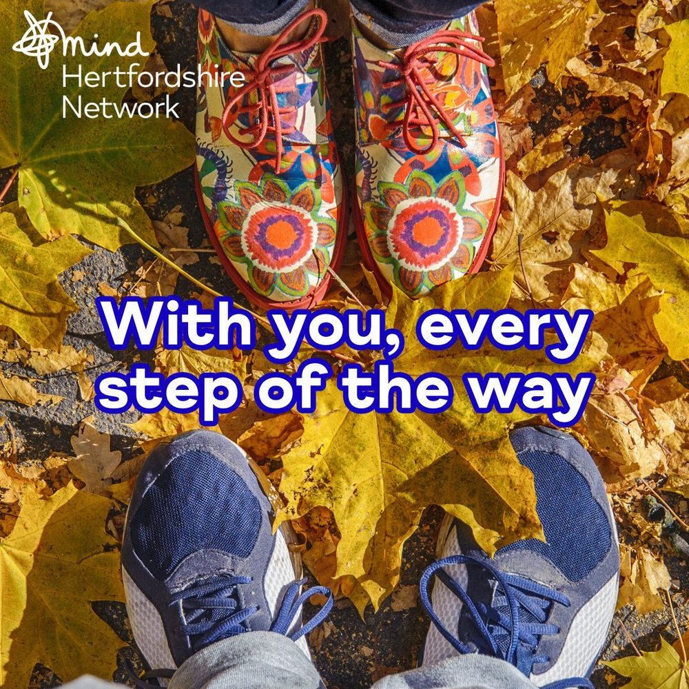 Two pairs of shoes are seen on a ground covered with autumn leaves. One pair is colourful with floral patterns, and the other is blue and white. The text reads, “With you, every step of the way”. The Hertfordshire Mind Network logo is in the top left corner