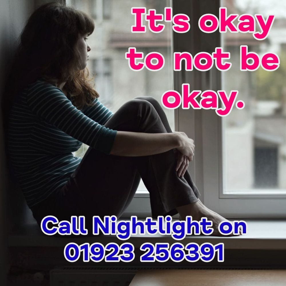 A woman sits on a window ledge, looking outside with a thoughtful expression. The text reads, “It’s okay to not be okay.” Below, it says, “Call Nightlight on 01923 256391.”