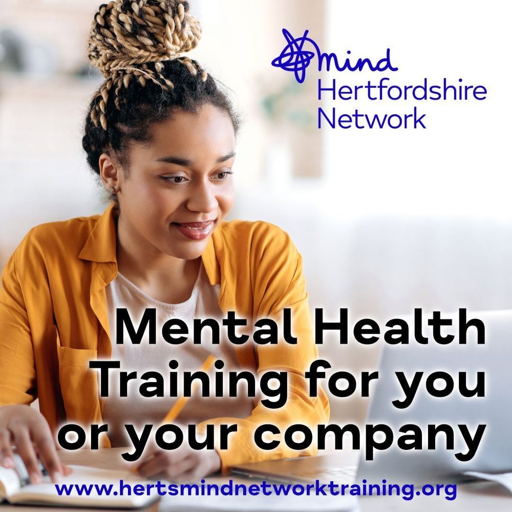 A smiling woman with braided hair sits at a desk, writing in a notebook while looking at a laptop screen. In the top right corner, the logo for ‘Mind Hertfordshire Network’ is displayed in blue. Bold black text in the foreground reads: ‘Mental Health Training for you or your company.’ Below, the website address ‘www.hertsmindnetworktraining.org’ is shown in blue text.
