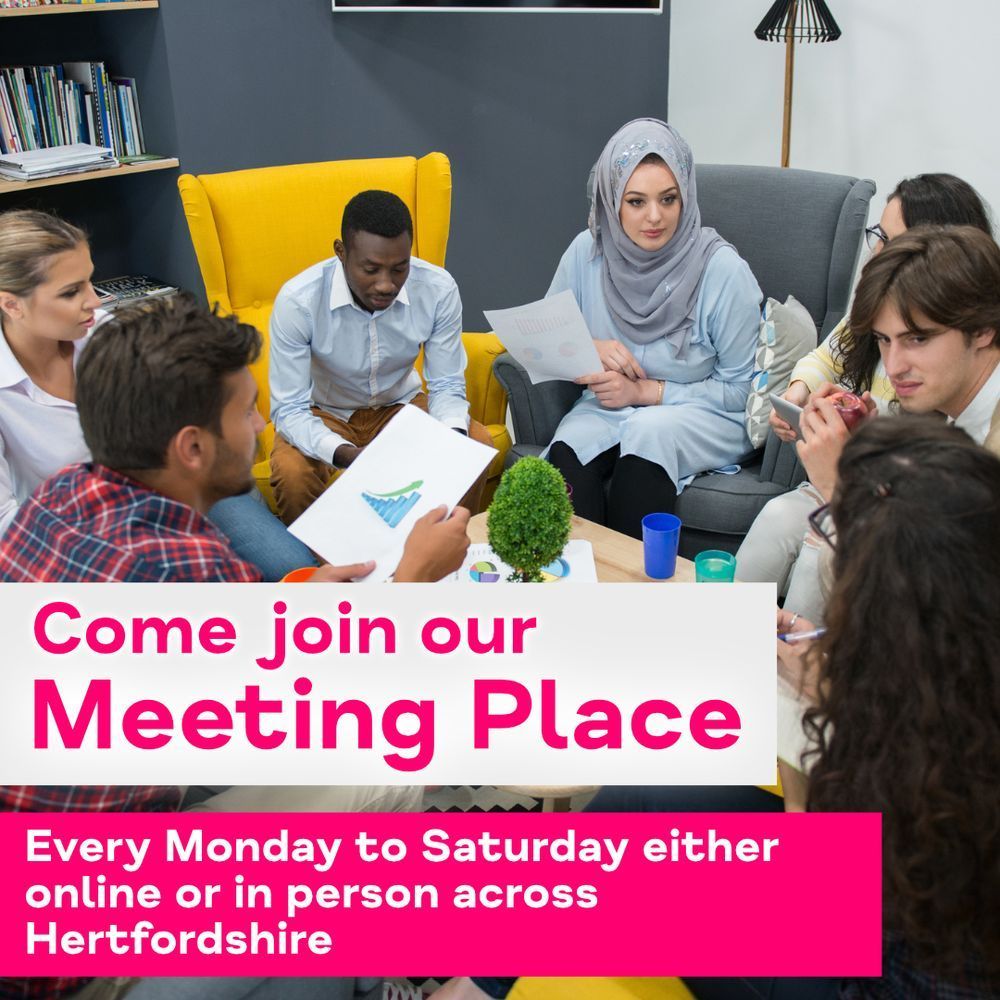 A group of 7 individuals sitting in a circle around a table with looking at some papers and appearing in conversation. The text reads 'Come join our Meeting Place, Every Monday to Saturday either online or in person across Hertfordshire'