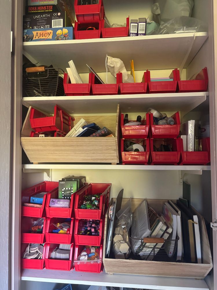 Red plastic bins filled with various board game bits and bobs (and some office supplies) in a small storage closet.