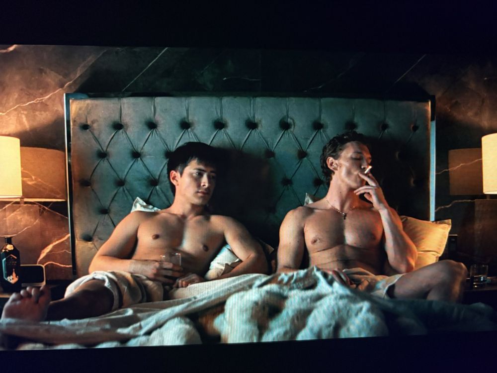Two men sit naked in a rumpled bed, one is holding a glass and the other is smoking a cigarette. The lighting is moody and langorous.