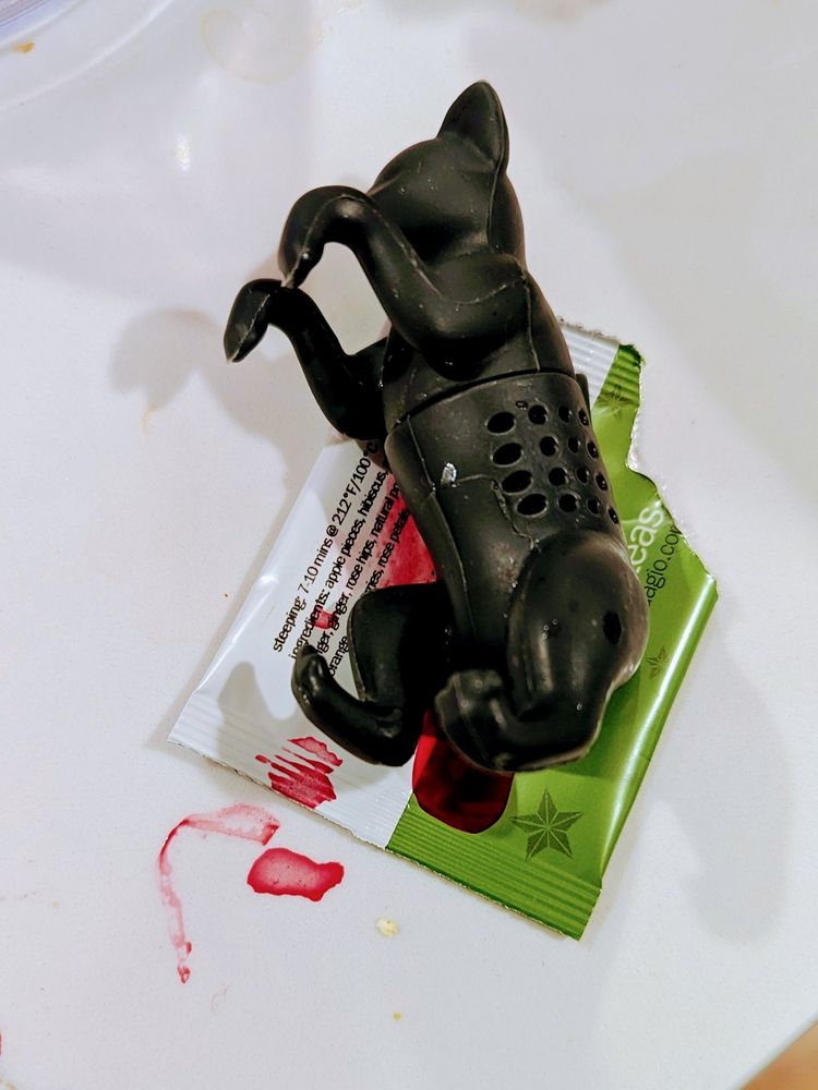 A black tea steeper shaped like a cat with its front paws up, rests on a green and white tea sachet. A red liquid has seeped out of the holes in the cat and pooles under it. The liquid is tea, but looks, horrifyingly, kind of like blood.