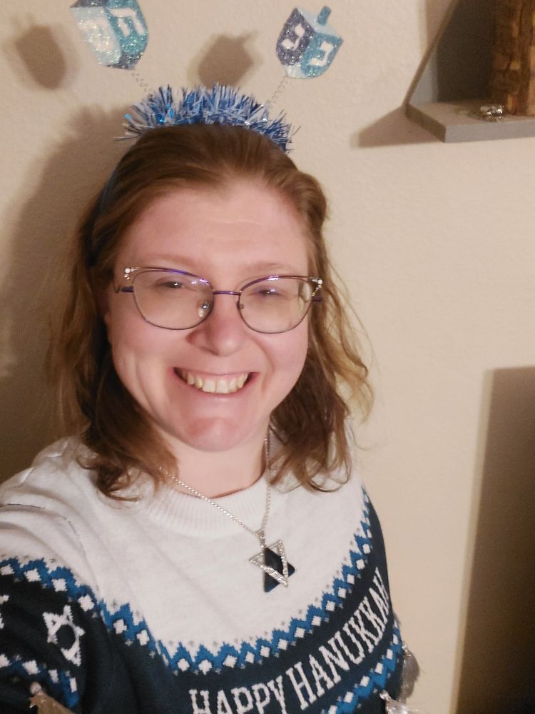 Woman wearing a sweater that says "Happy Hanukkah", a large star of David, and a headband with dreidels.