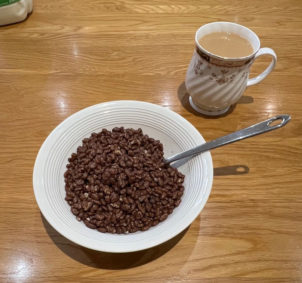 A bowl of Coco Pops and a cup of tea with milk on a wooden kitchen table. 