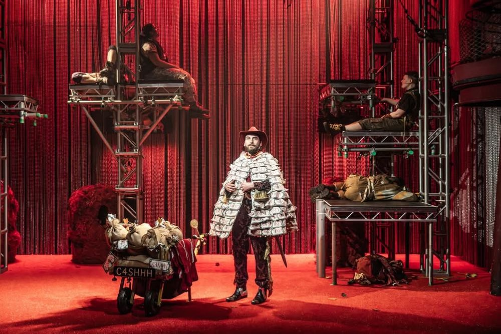 A picture of a theatre stage. The floor is a texture red. There is a silver chain mail backdrop which appears red under the lights. There are two scaffolding structures on either side of the image with platforms attached partway up. A man stands on the ground next to a wheeled cart. He is wearing a cape covered in money along with a red cowboy hat. On the platforms there are three other figures who appear to be asleep. They are wearing fleeces and green or khaki trousers.