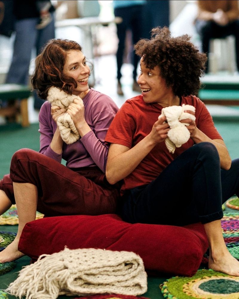 Two women sit back to back on a large red cushion but are turned to look at each other. They are both clutching white bobble hats and looking excited. In front of the cushion is a white knitted scarf, folded over on itself. Surrounded the cushion are circular fabric mats made of knotted green, yellow and red material.