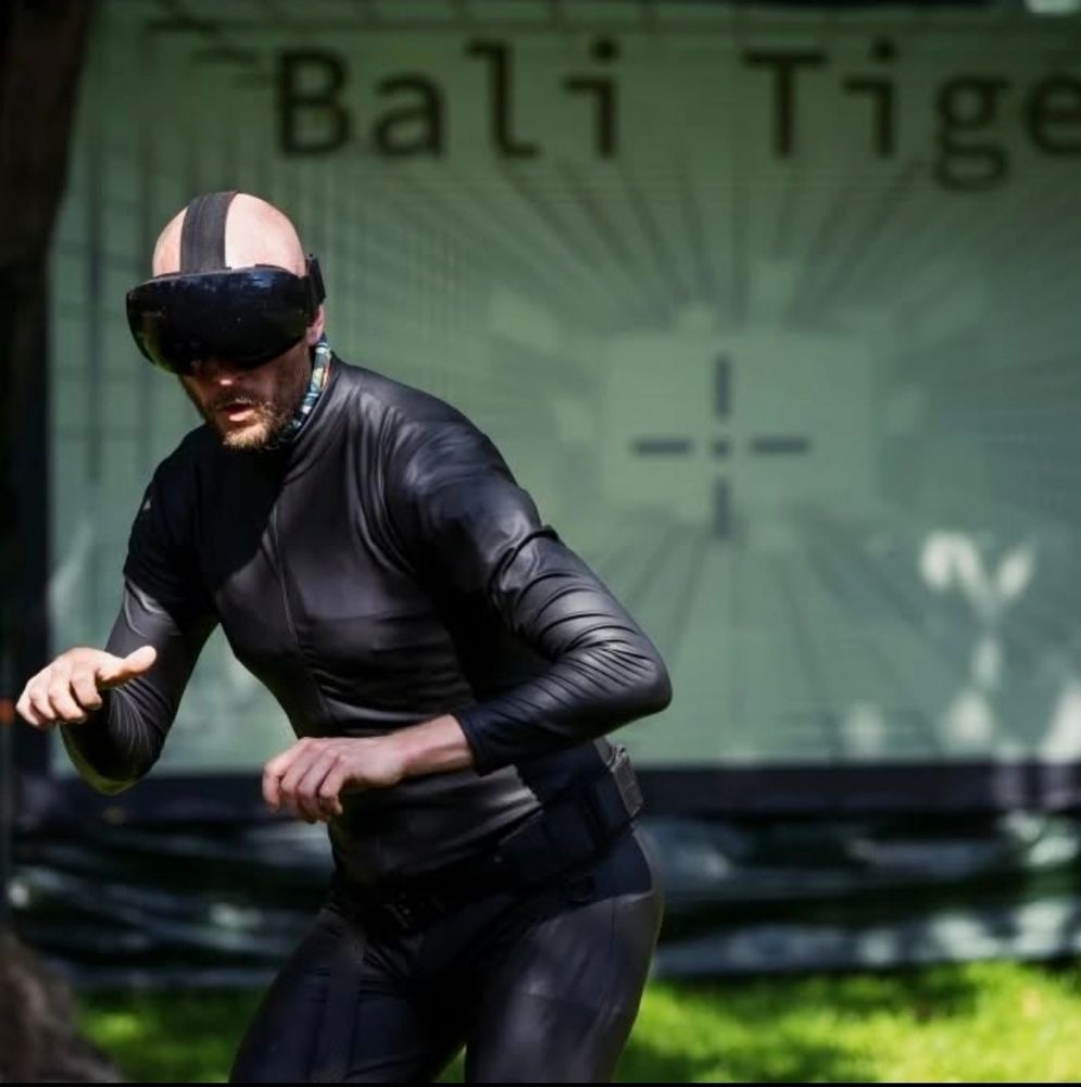 A bald man wearing a black wetsuit and black VR headset is slightly crouched on the left. Behind him is a large screen with the words "bali tiger" on. There is grass and some shadows cast on the screen.