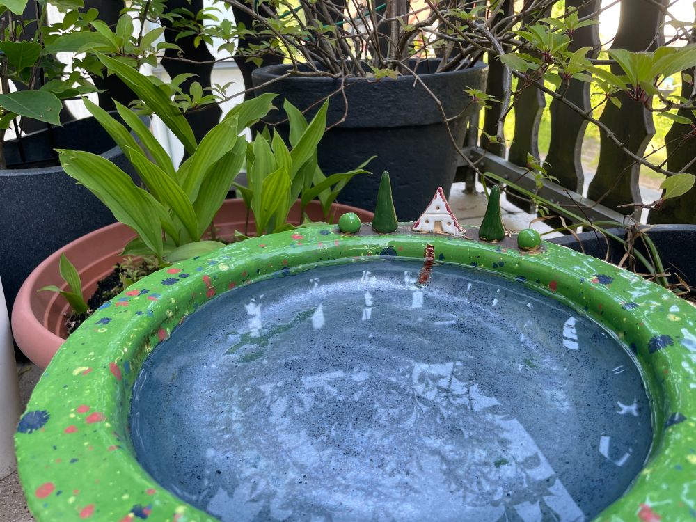 Handgefertigte runde Schale, innen blau-silber (Wasser), der Rand grün mit bunten Punkten ( Blumenwiese) lackiert. Auf dem Rand steht ein kleines weißes Häuschen mit rotem Dach, daneben jeweils eine dunkelgrüne hohe Tanne und ein kleiner kugelförmiger, hellgrüner Laubbaum. Vor dem Haus führt eine kleine braune Leiter von der Wiese hinab ins Wasserund lädt zum Baden ein. 