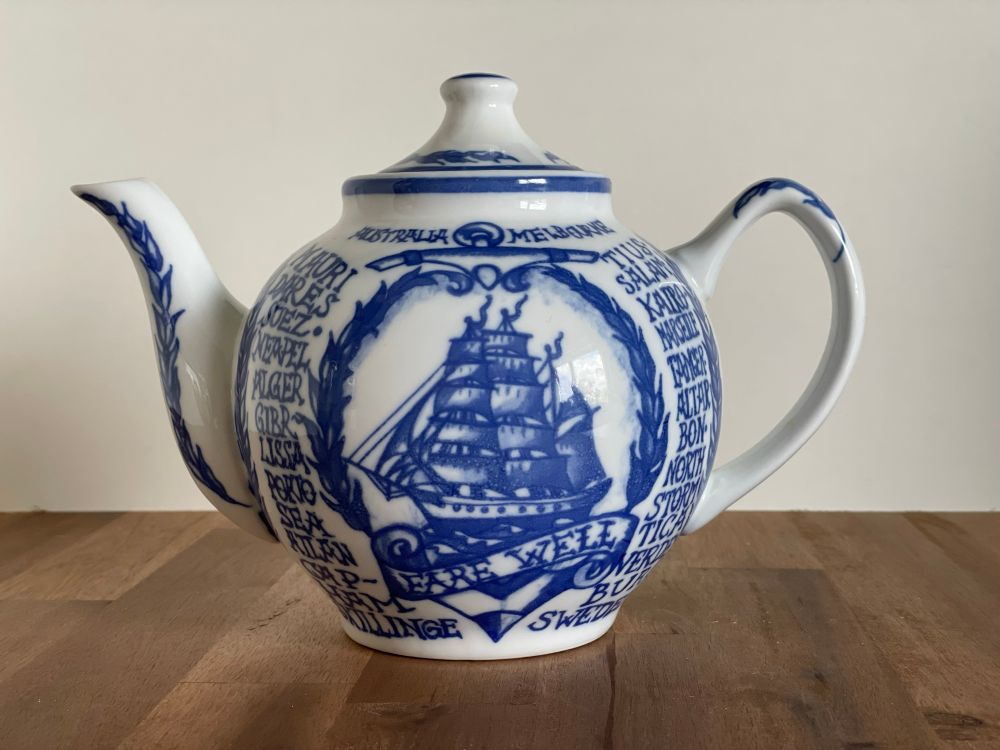 Bauchige, weiße Porzellan-Teekanne mit blauer Bemalung und Schrift. Mittig ist ein blaues Segelschiff, darunter der Schriftzug „fare well“. Das Schiff war unterwegs auf einer Reise von Schweden nach Australien und wieder zurück. Auf dem Rest der Kanne, um das Schiff herum, kann man die verschiedenen Häfen nachlesen, die es angelaufen hat. 