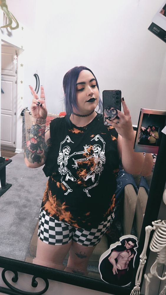 a mirror selfie of a girl in a tye dye black and orange shirt either dancing skeletons on it and black and white checkered shorts 