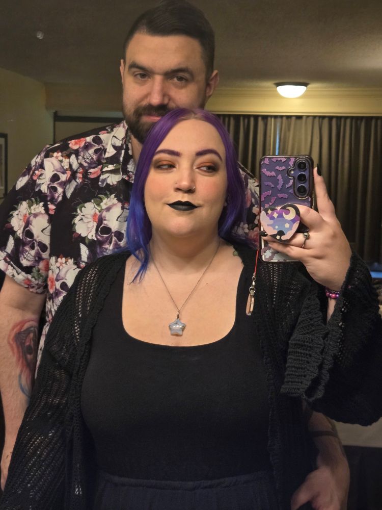 a mirror selfie of a girl with purple hair in a black shirt with a man with a dark hair and beard behind her