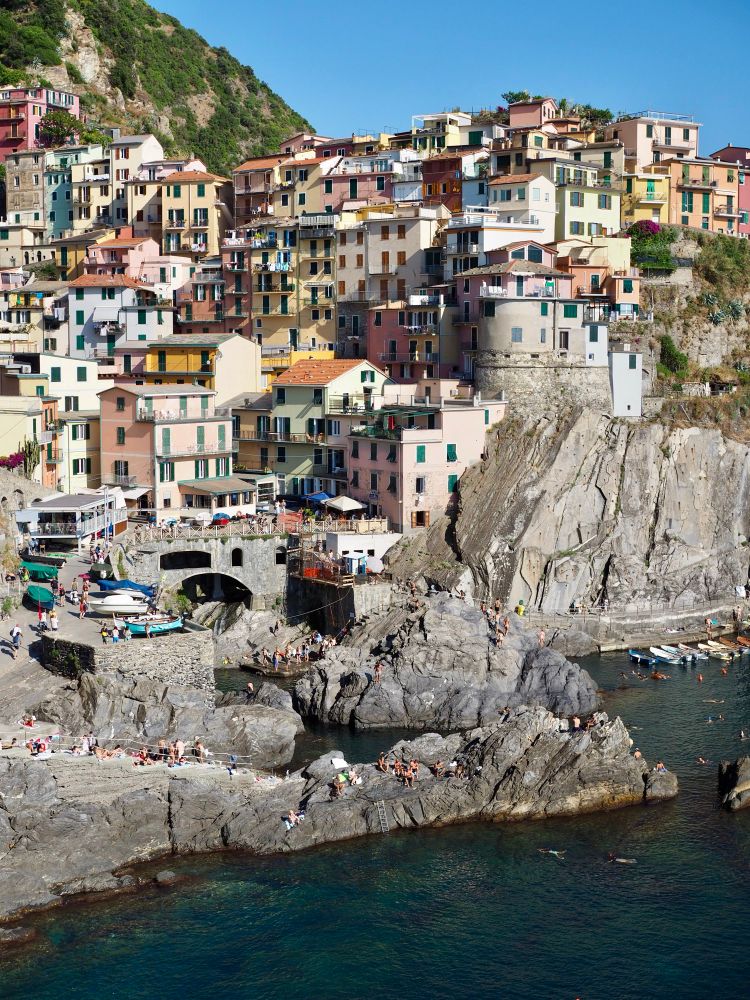 イタリア、マナローラの景色。崖の上から下にかけてカラフルな家が立ち並び、一番下の岩場には飛び込みや日光浴を楽しむ人々が見える。
