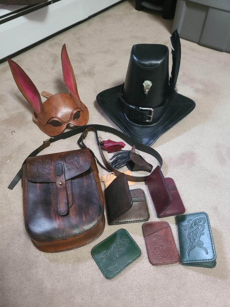 A lot of leather items on the floor: wallets, key fogs, a raven bag, a bunny mask and a witch hunter hat.