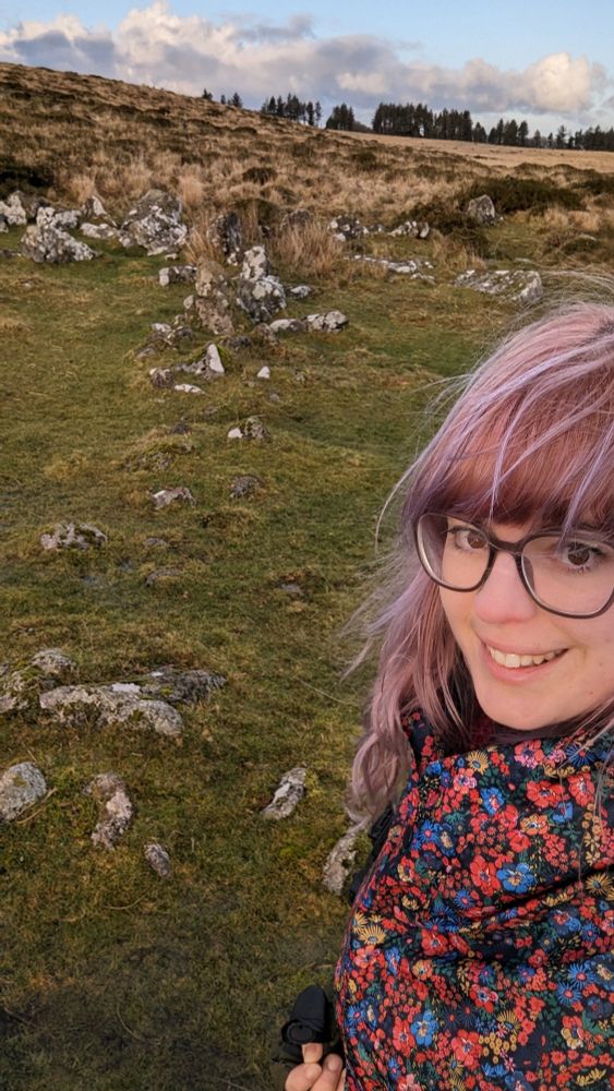 A line of collapsed bronze age granite wall with grassy moorland in the background, and a girl with pink hair and red flowery jacket in the foreground