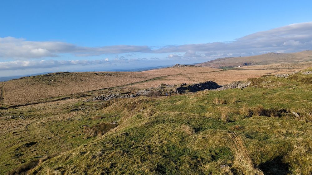 A moorland view, grassy and mossy, with a cloudy and also blue sky , some rocky tors in the distance and two people walking