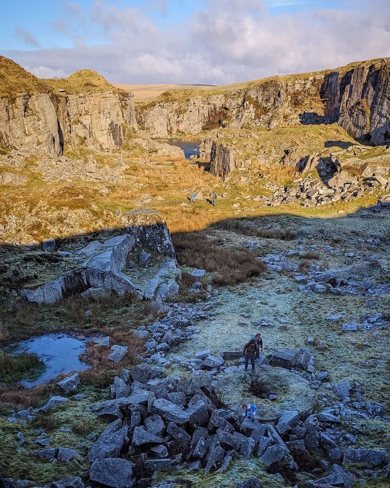 Foggintor quarry, a large rocky dip with the sun shining on one half, frosty grass in the shade, and three people that look very tiny about to walk up a steep hill where the photographer is standing