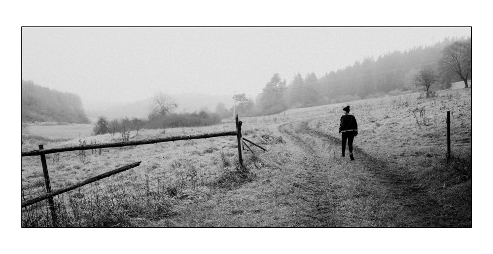 A black-and-white photograph of a wintery rural landscape with frosted grass, rolling hills, and a faint tree line fading into mist. A person wearing a jacket and hat walks alone along a winding dirt path toward the right side of the frame. A simple wooden fence stretches diagonally across the foreground.
